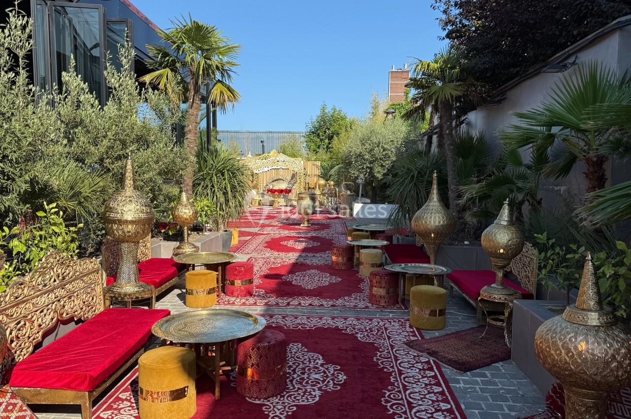 Allée décorée de tapis rouges, mobilier oriental doré et végétation luxuriante dans un cadre extérieur ensoleillé.