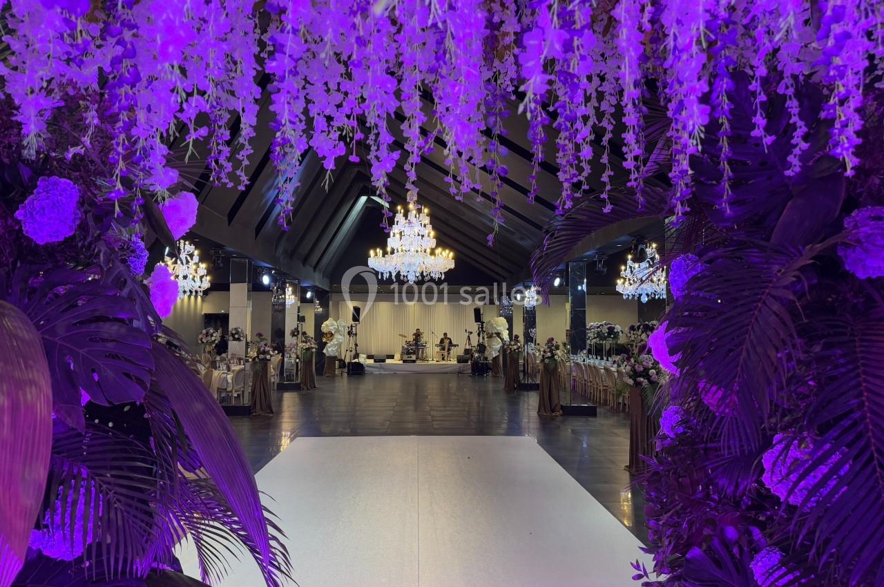 Salle de réception décorée de guirlandes florales violettes, avec chandeliers suspendus et tables dressées.