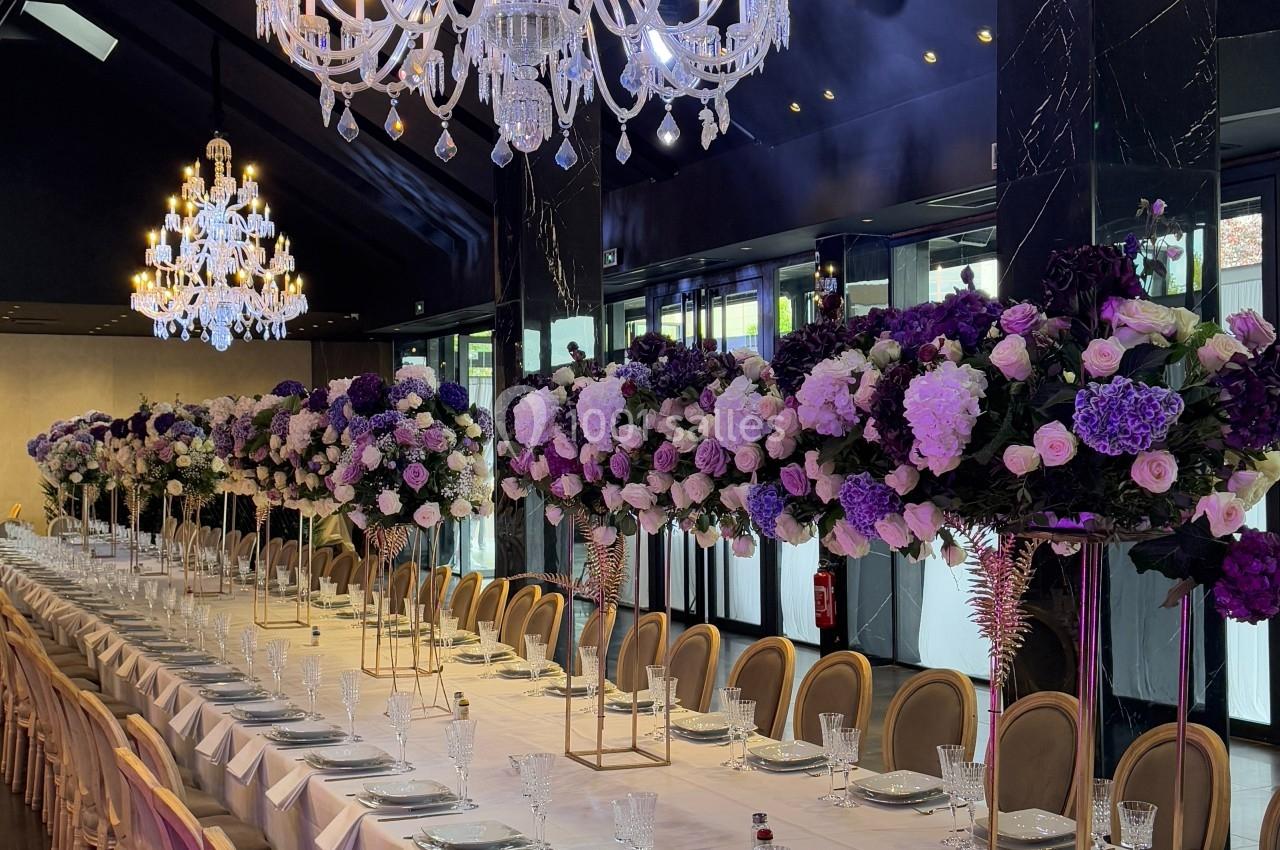 Salle de réception élégante avec longues tables dressées, chandeliers en cristal et décorations florales violettes.