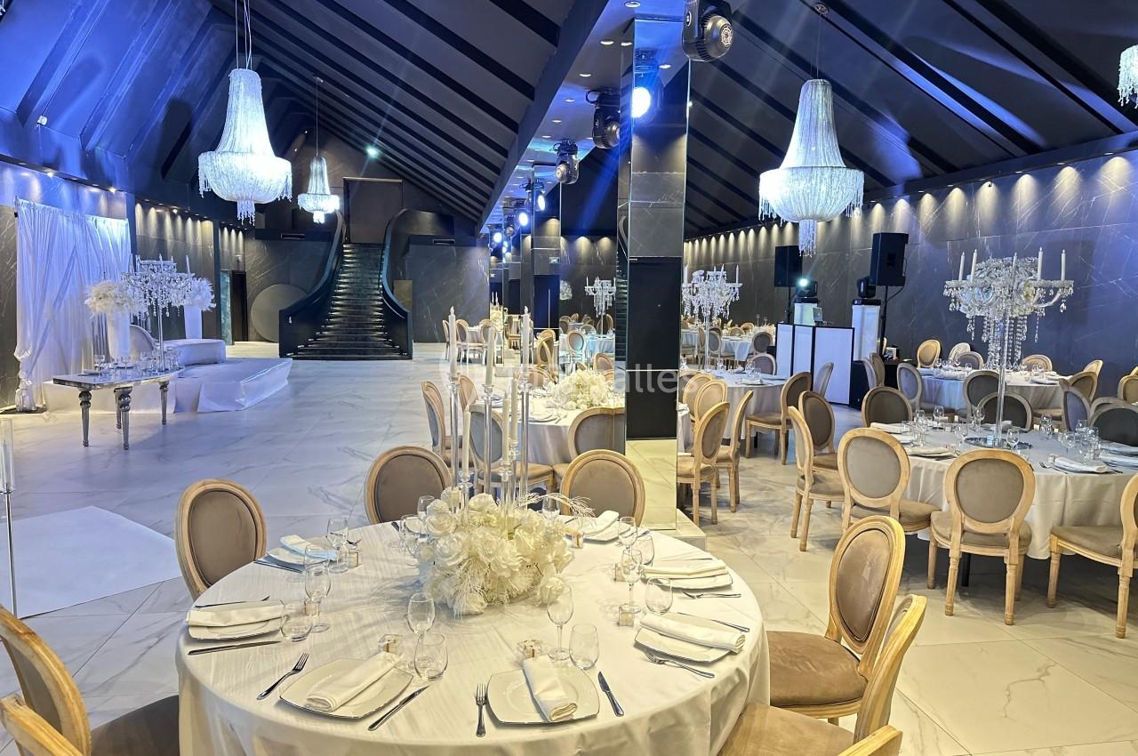 Salle de réception élégante avec tables rondes dressées, chandeliers et décorations lumineuses sous un plafond en pente.