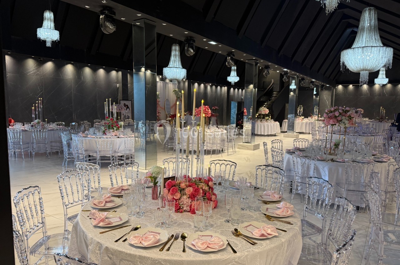 Salle de réception élégante avec tables décorées de nappes blanches, chandeliers et arrangements floraux roses.