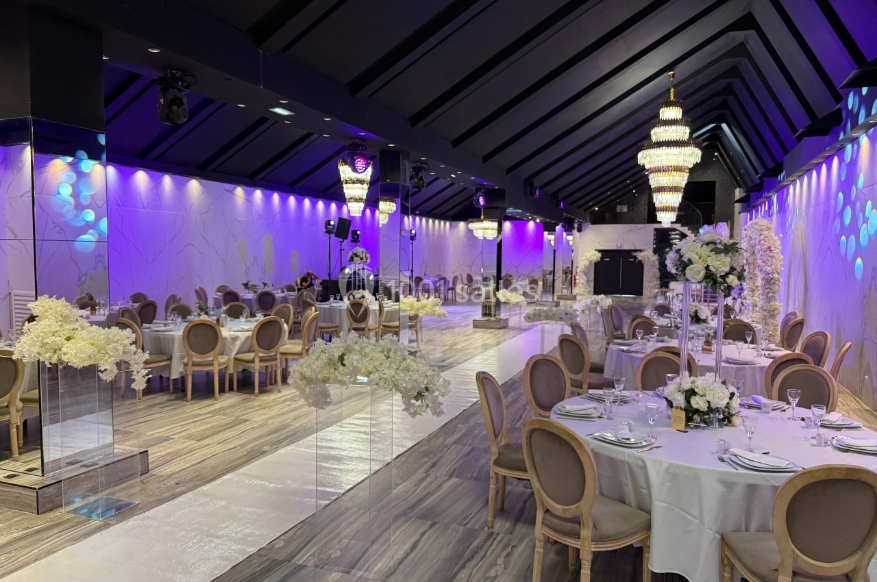 Salle de réception élégante avec tables dressées, décor floral blanc et lustres imposants sous un éclairage violet.