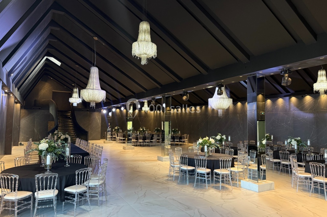 Salle de réception élégante avec tables rondes dressées, chaises transparentes et lustres suspendus.