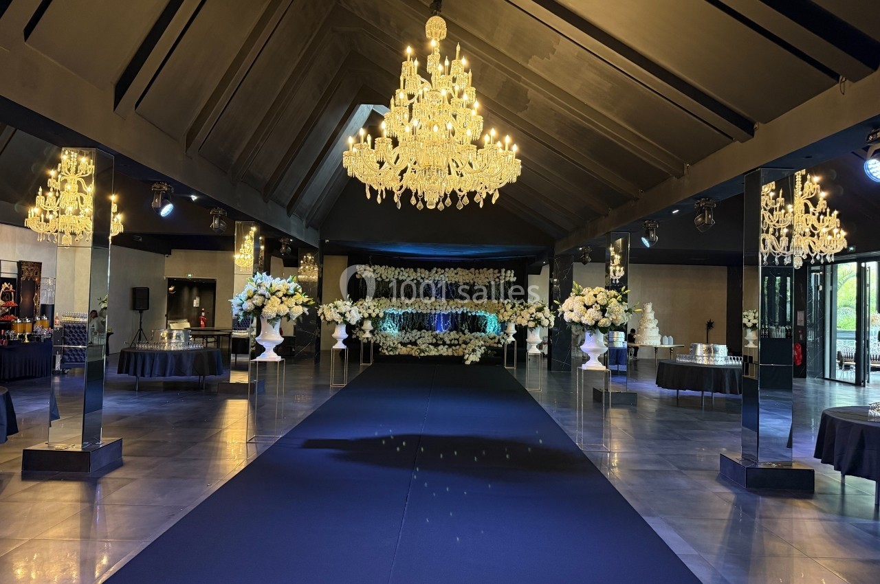 Salle de réception élégante avec lustres imposants, allée centrale noire et décorations florales blanches.
