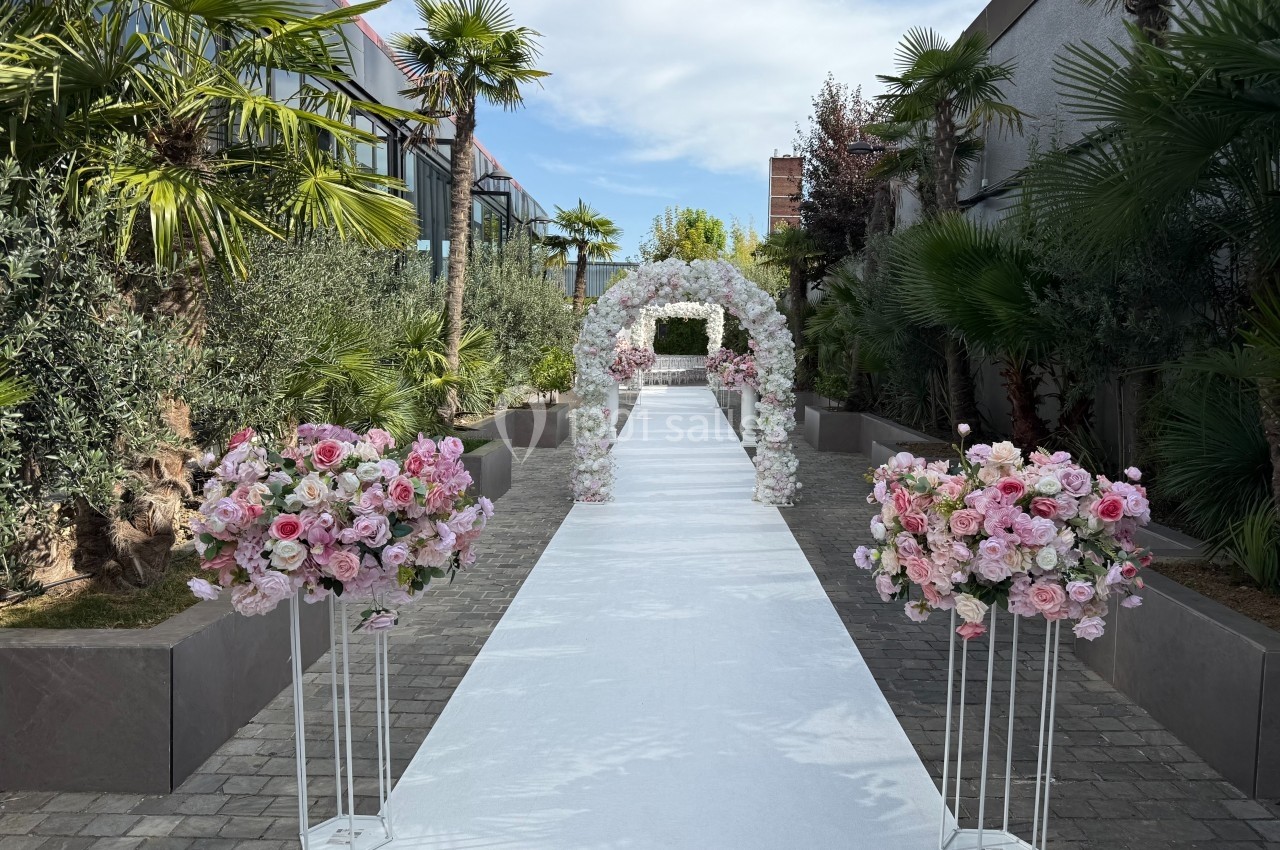 Allée extérieure décorée pour un mariage, avec arche florale blanche et arrangements de fleurs roses sur supports.
