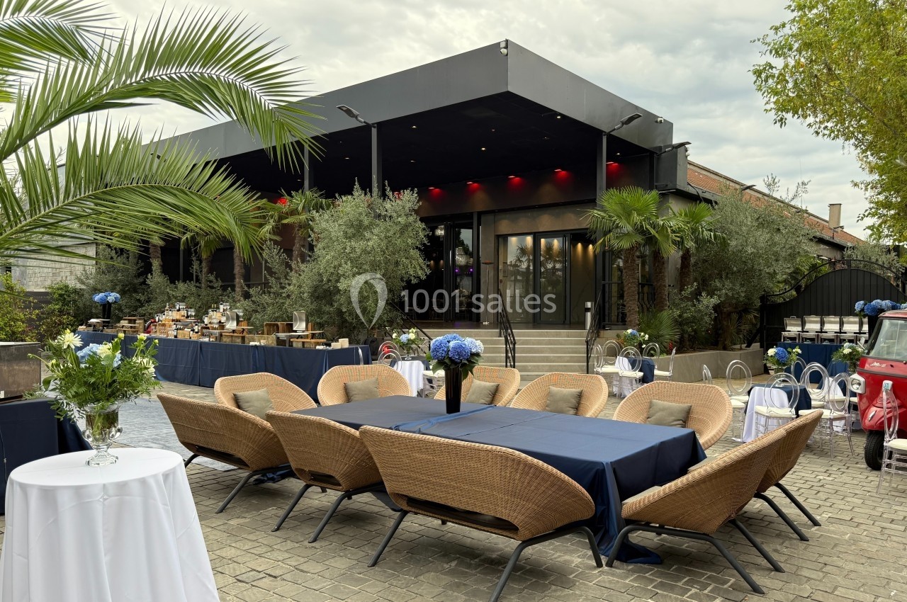 Terrasse aménagée avec des tables recouvertes de nappes bleues, entourées de fauteuils en rotin, devant un bâtiment moderne.