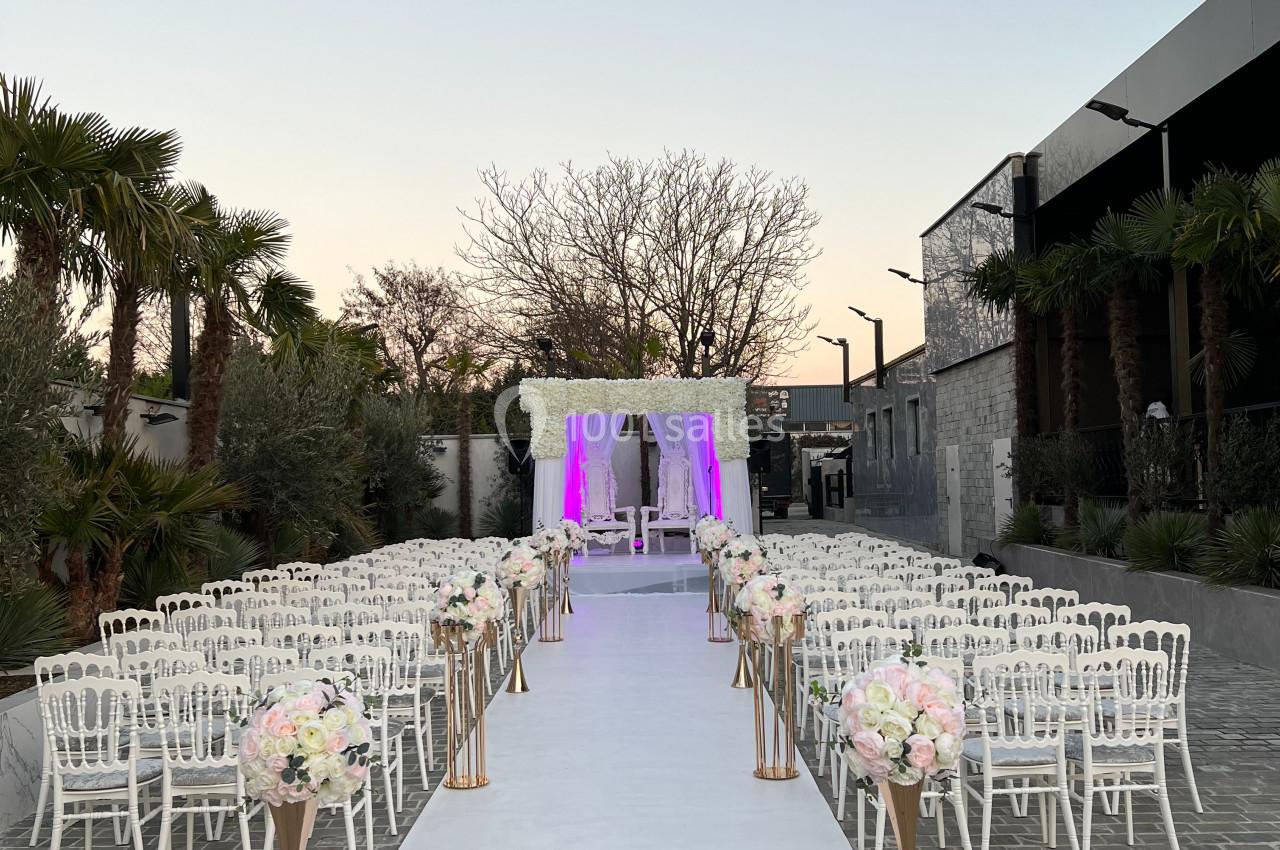 Allée centrale décorée de fleurs menant à une arche de mariage illuminée, entourée de chaises blanches en extérieur.