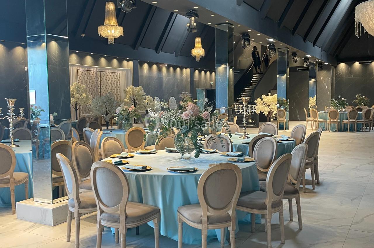 Salle de réception élégante avec tables rondes dressées, nappes bleues, chaises en bois clair et décorations florales.