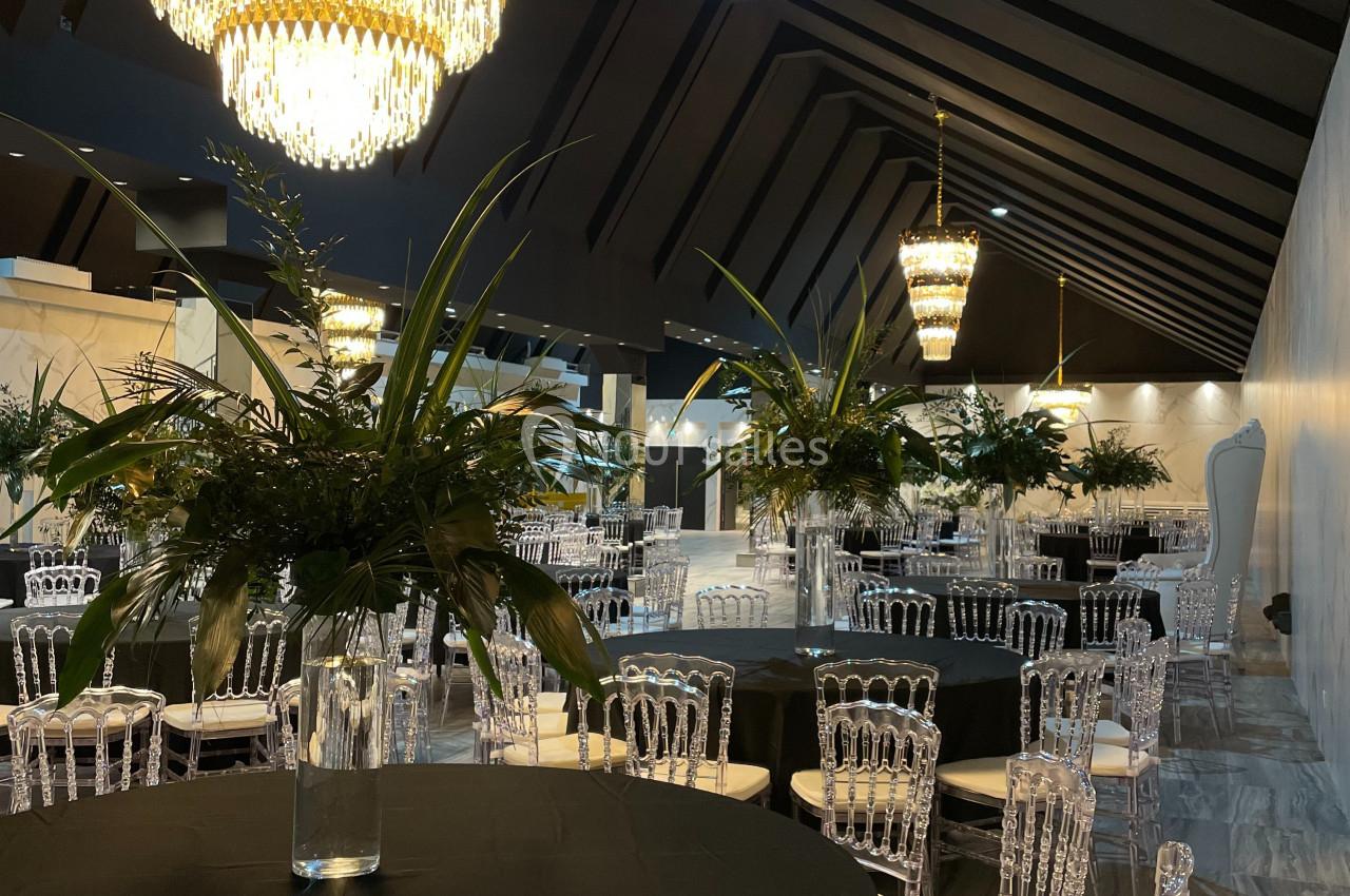 Salle de réception élégante avec tables rondes noires, chaises transparentes, lustres en cristal et décorations végétales.