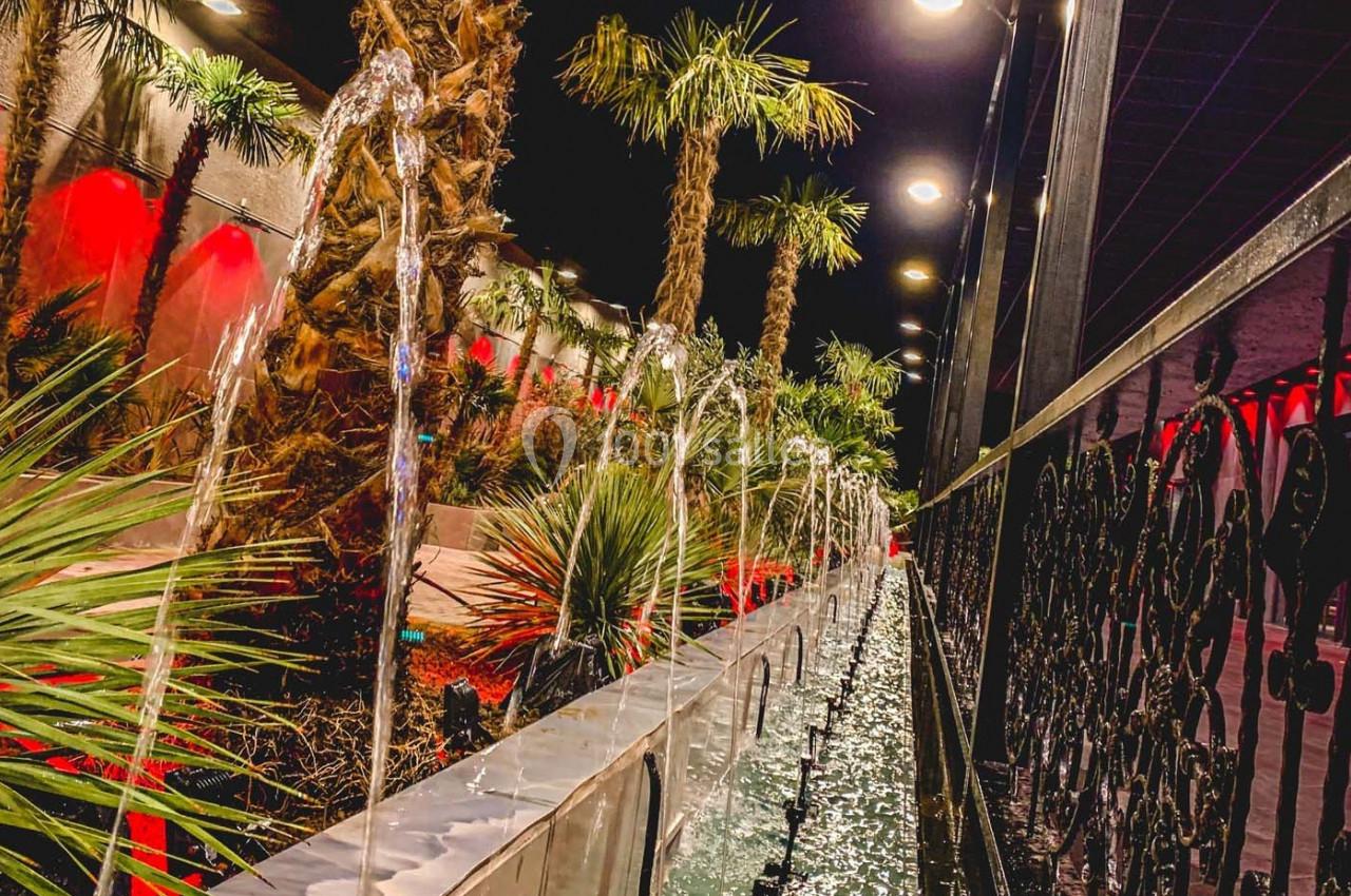 Allée bordée de palmiers et d'une fontaine avec jets d'eau, éclairée par des lumières rouges et jaunes.