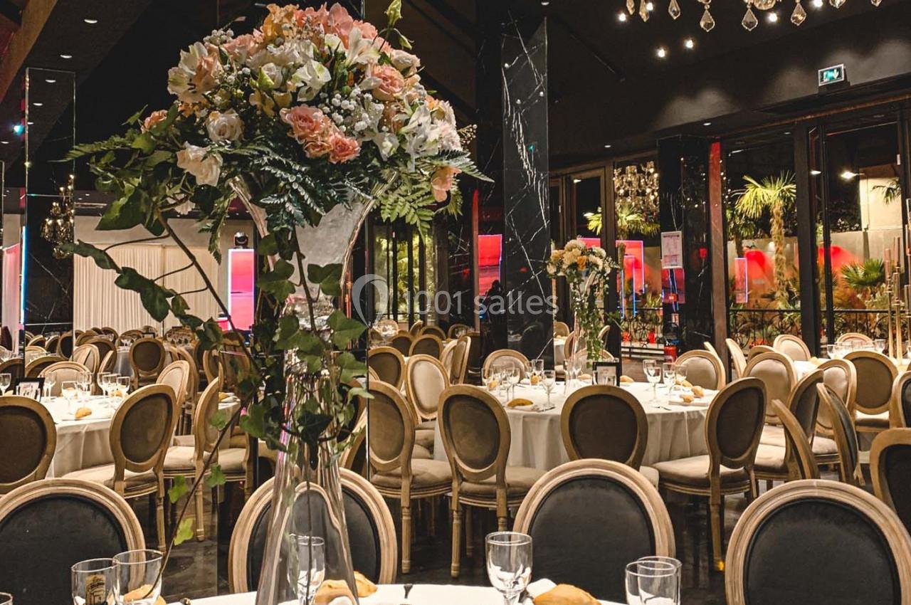 Salle de réception élégante avec tables rondes dressées, chaises en bois et grand bouquet floral au premier plan.
