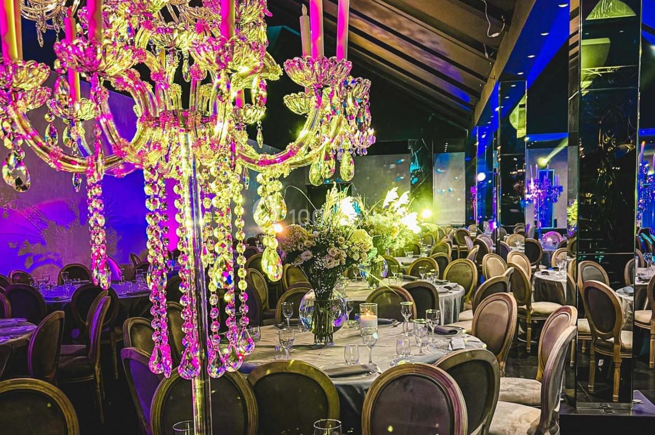 Salle de réception élégante avec lustre en cristal, tables décorées de fleurs et éclairage coloré.