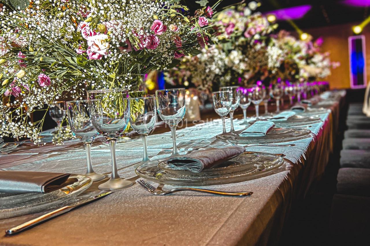 Table élégamment dressée avec nappes beige, verres à pied, couverts et bouquets de fleurs blanches et roses.