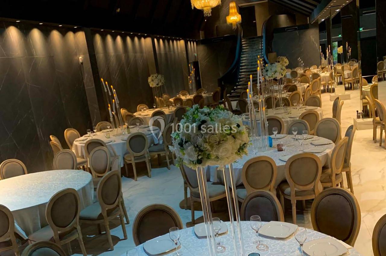 Salle de réception élégante avec tables rondes dressées, chaises en bois clair et décorations florales blanches.