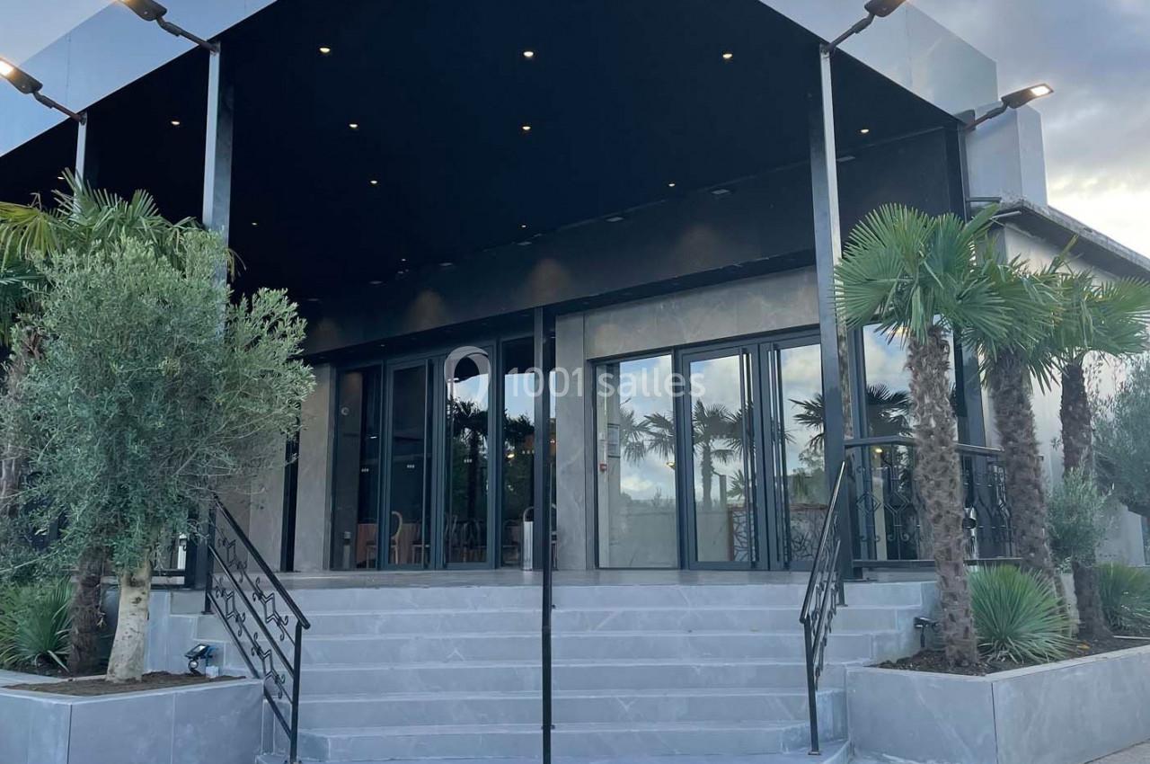 Entrée d'un bâtiment moderne avec des escaliers, des portes vitrées, des palmiers et des éclairages extérieurs.