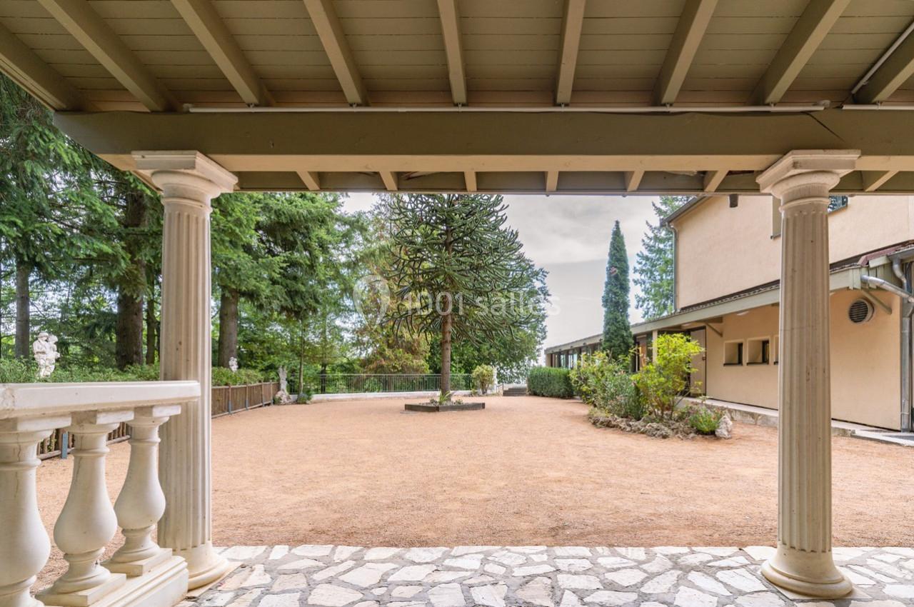 Vue d'une cour extérieure avec sol en gravier, entourée de végétation et partiellement abritée par une terrasse à colonnes.