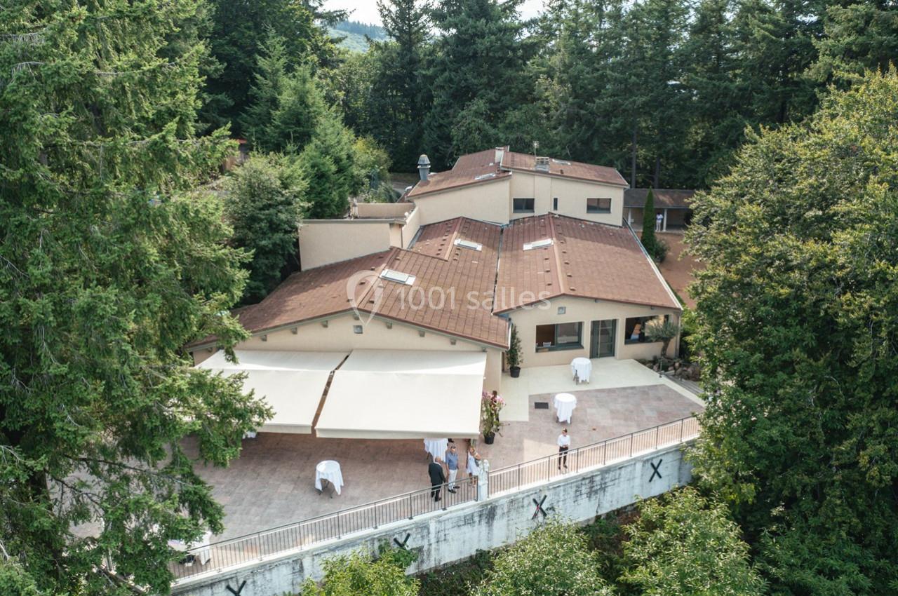 Vue aérienne d'un bâtiment entouré de végétation, avec une terrasse aménagée et des personnes présentes.