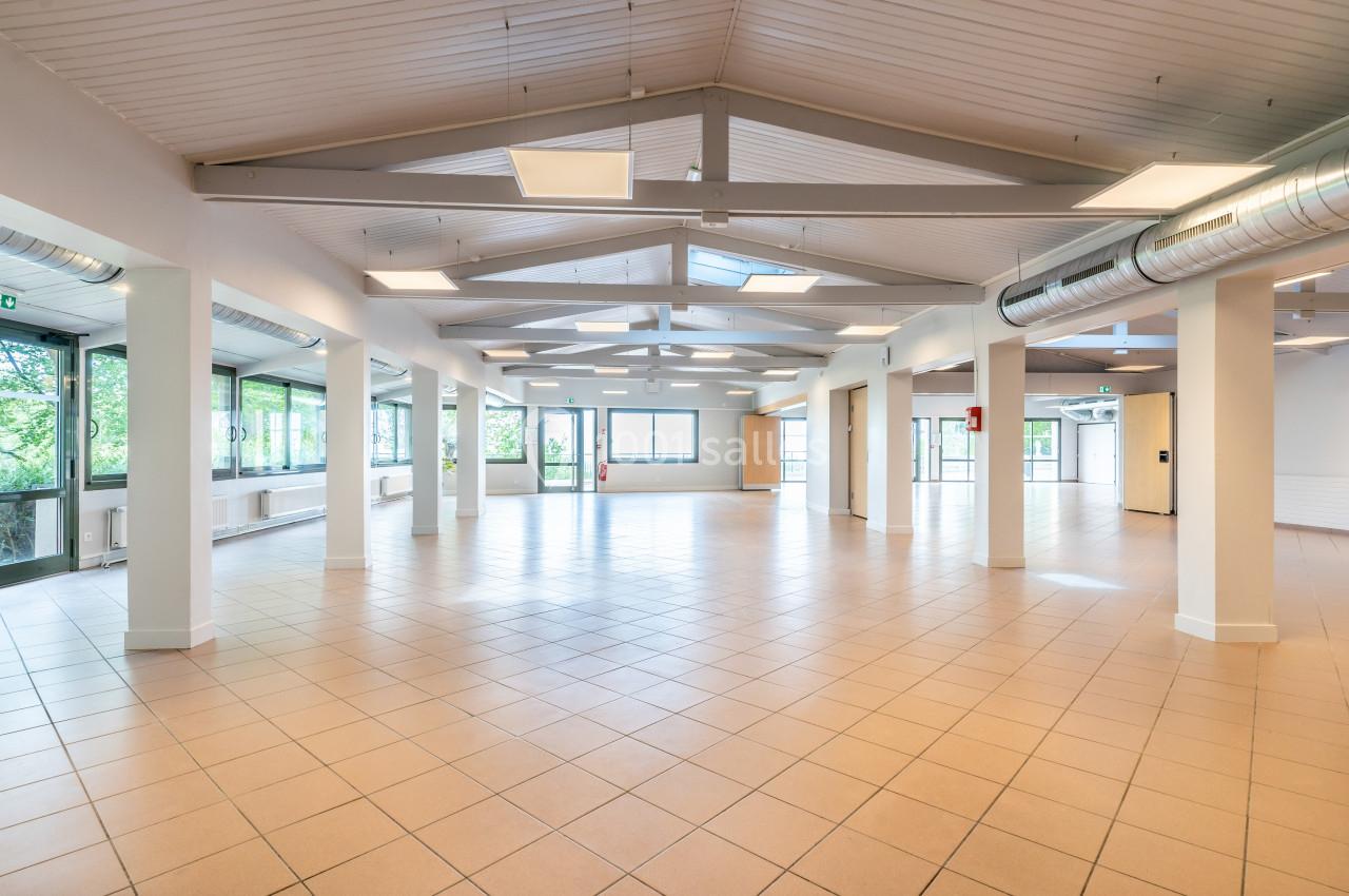 Salle spacieuse avec carrelage au sol, plafond en poutres apparentes et grandes fenêtres laissant entrer la lumière…