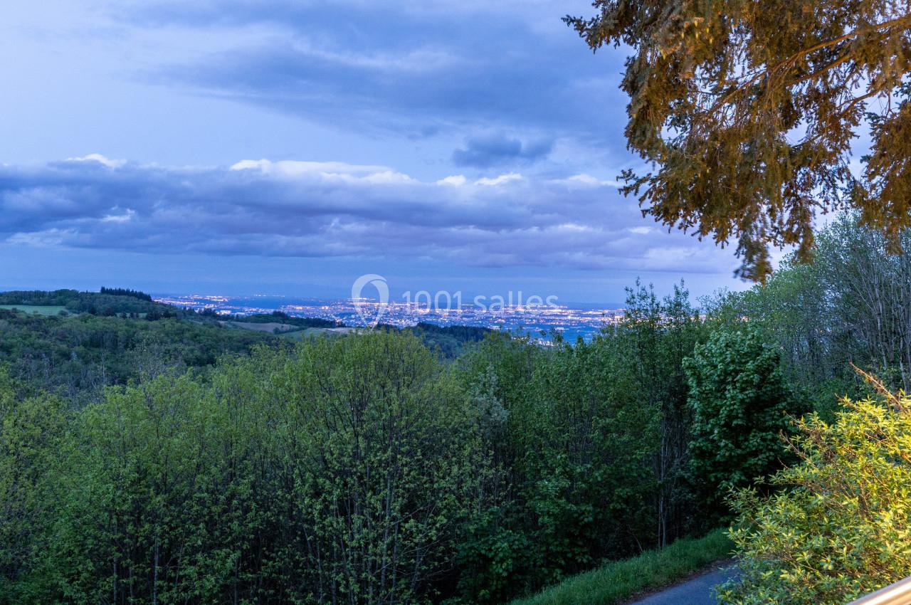 Vue nocturne d'une ville illuminée à l'horizon, entourée de collines boisées sous un ciel nuageux.