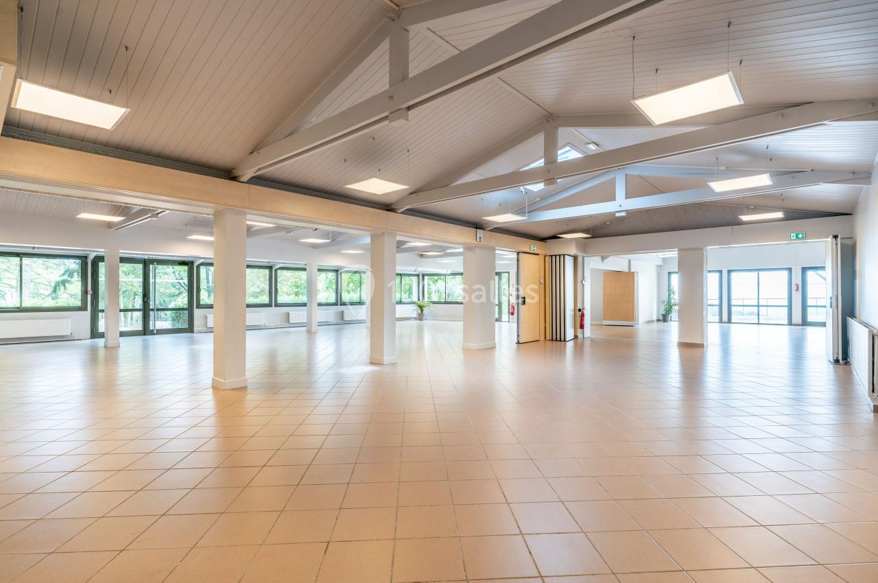 Salle lumineuse avec plafond en poutres apparentes, carrelage beige et grandes fenêtres donnant sur un espace verdoyant.