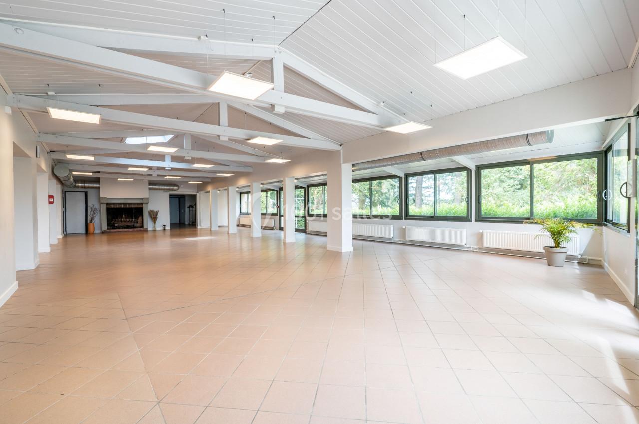 Salle spacieuse et lumineuse avec un sol carrelé, de grandes fenêtres et un plafond en poutres blanches.
