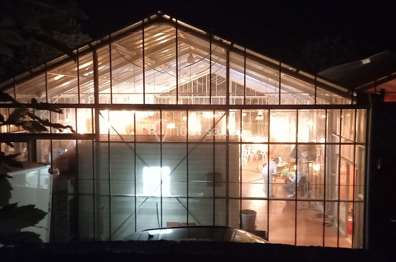 Vue nocturne d'une serre éclairée de l'intérieur, révélant des activités et des équipements visibles à travers les vitres.