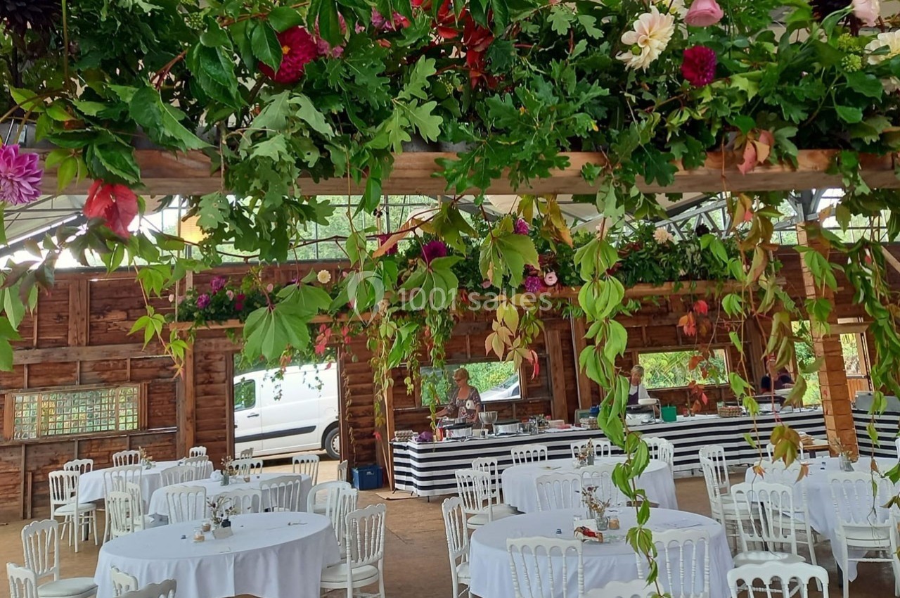 Salle décorée pour un événement avec tables rondes, chaises blanches et guirlandes de fleurs suspendues.
