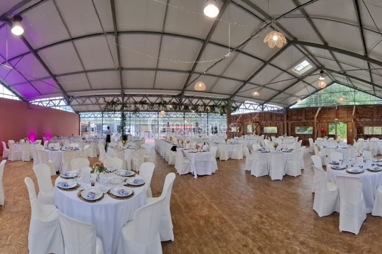 Salle de réception spacieuse avec tables rondes dressées, nappes blanches et décorations suspendues sous une structure…