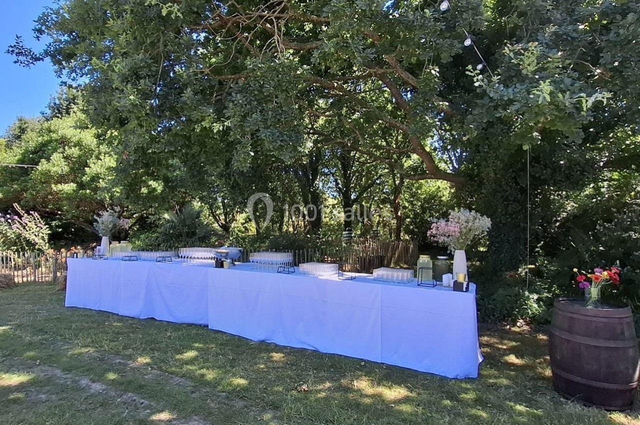 Table dressée avec nappes blanches en extérieur, sous des arbres, décorée de fleurs et entourée de verdure.