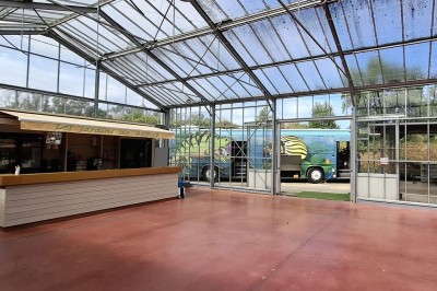 Miniature Location salle Névez (Finistère) - Jardins et Serres de Rospico #12 Grande salle lumineuse avec sol rouge, toit en verre, chaises blanches, comptoir et décorations suspendues.