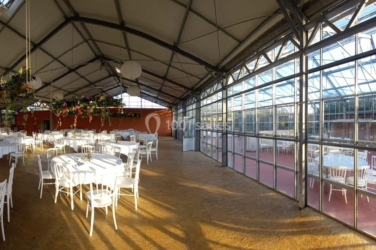 Salle lumineuse avec tables et chaises blanches, située dans une structure en verre et métal avec des plantes suspendues.