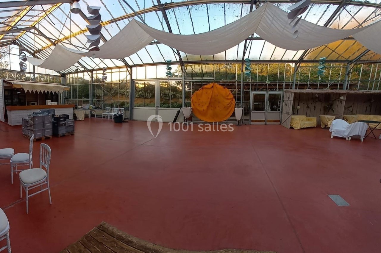 Grande salle lumineuse avec sol rouge, toit en verre, chaises blanches, comptoir et décorations suspendues.