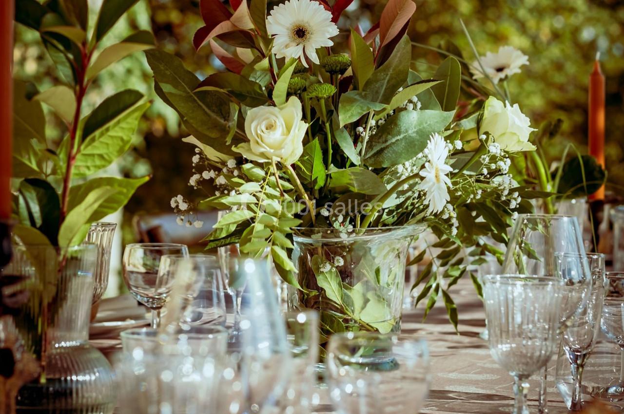 Centre de table floral avec roses blanches et feuillage, entouré de verres en cristal sur une table dressée en extérieur.