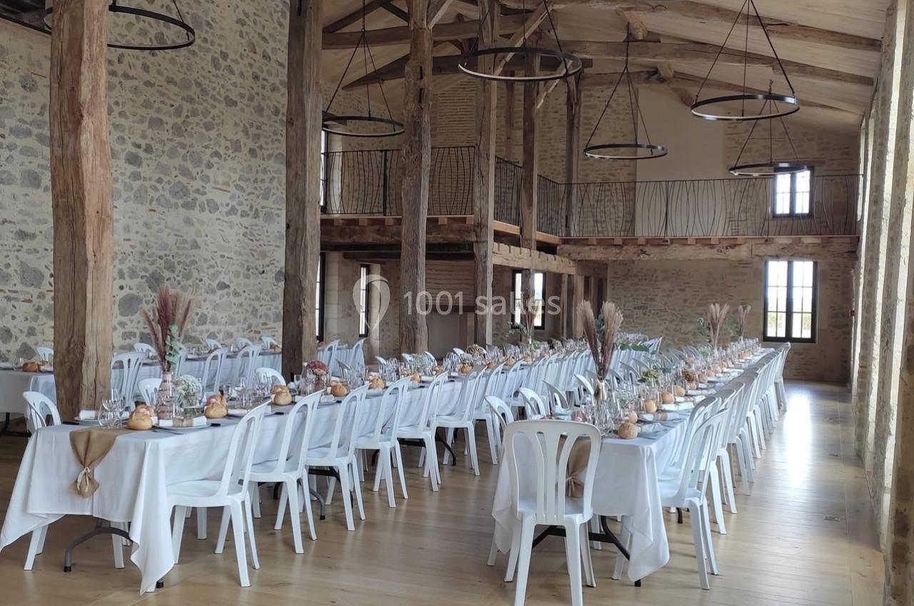 Salle de réception rustique avec tables alignées, nappes blanches, décorations florales et poutres apparentes en bois.
