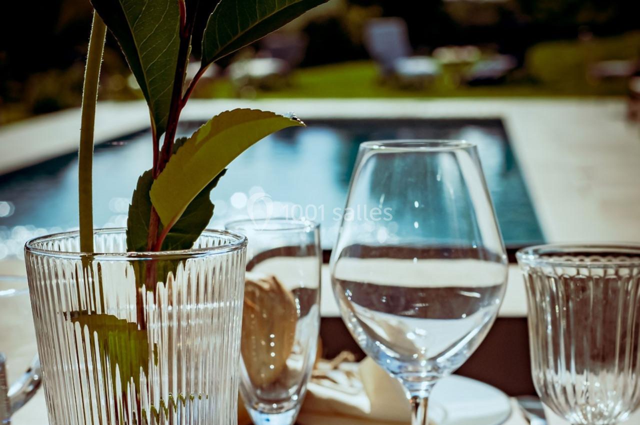 Verres et vase en verre sur une table extérieure, avec une piscine floue et un jardin en arrière-plan.
