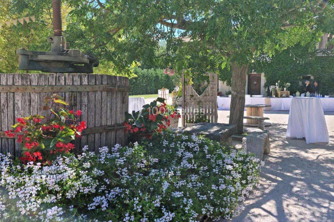 Jardin ombragé avec fleurs colorées, ancien pressoir en bois et tables dressées en arrière-plan.