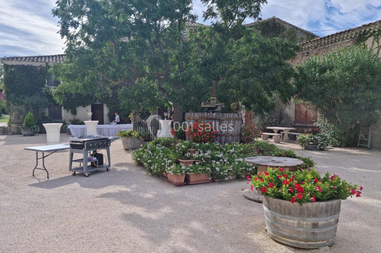 Cour extérieure avec arbres, fleurs en pots, tables et équipements disposés sur un sol en gravier devant un bâtiment en…