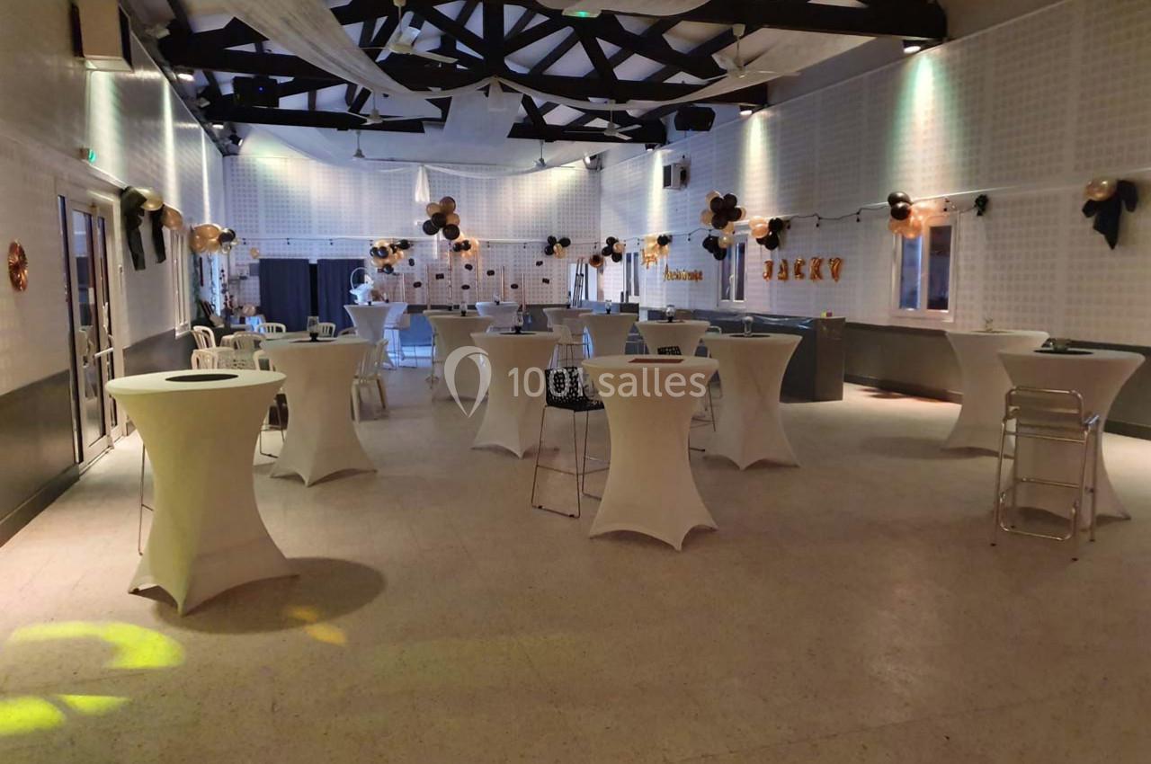 Salle décorée pour un événement, avec des tables hautes recouvertes de nappes blanches et des ballons suspendus.