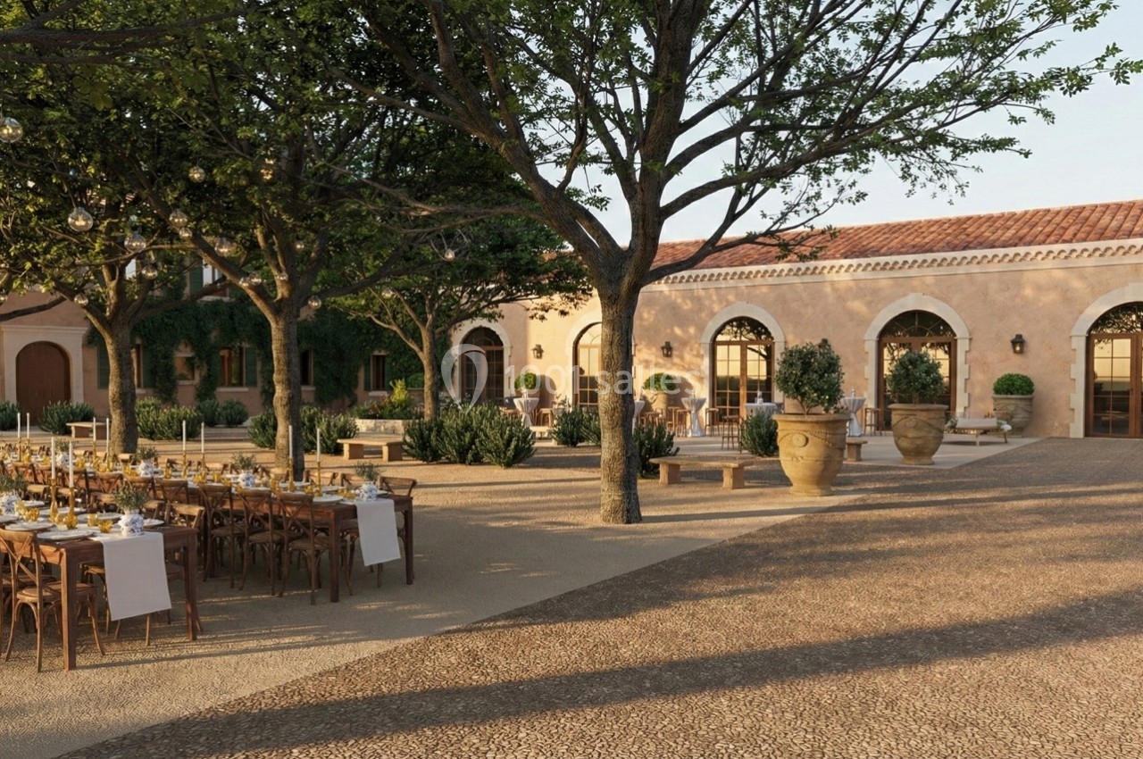Cour extérieure aménagée avec des tables dressées pour un repas, entourée d'arbres et de bâtiments aux arches en pierre.