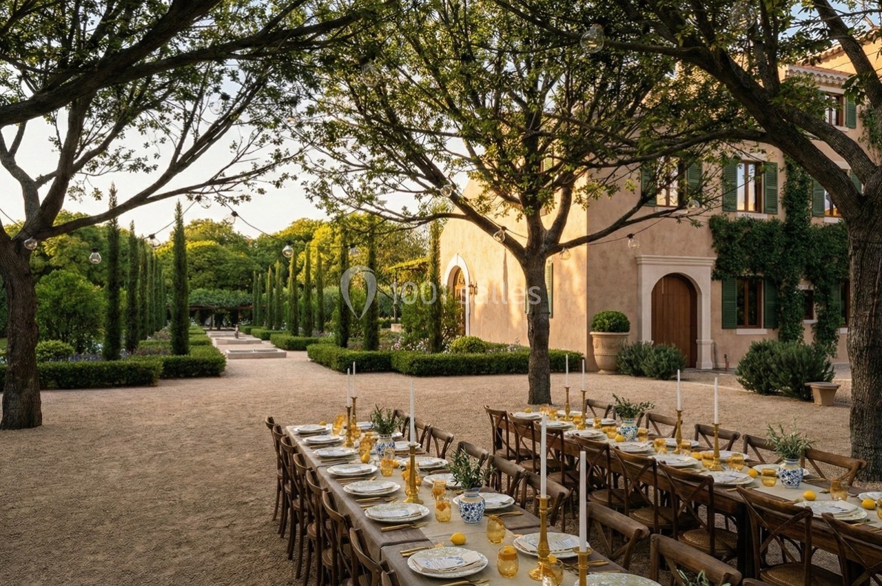 Grande table dressée en extérieur sous des arbres, avec vaisselle élégante, devant une maison entourée de verdure.