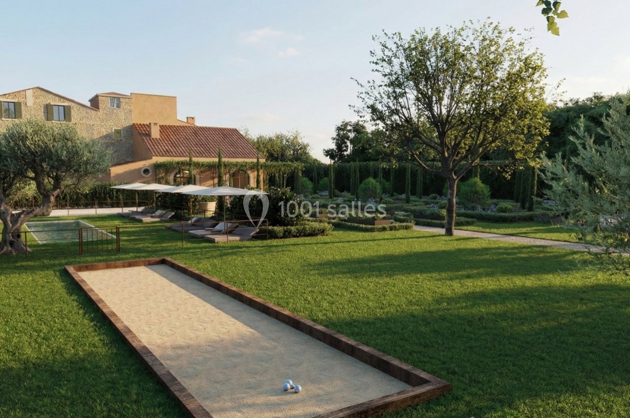Terrain de pétanque ensoleillé dans un jardin verdoyant avec une maison en pierre et des arbres en arrière-plan.