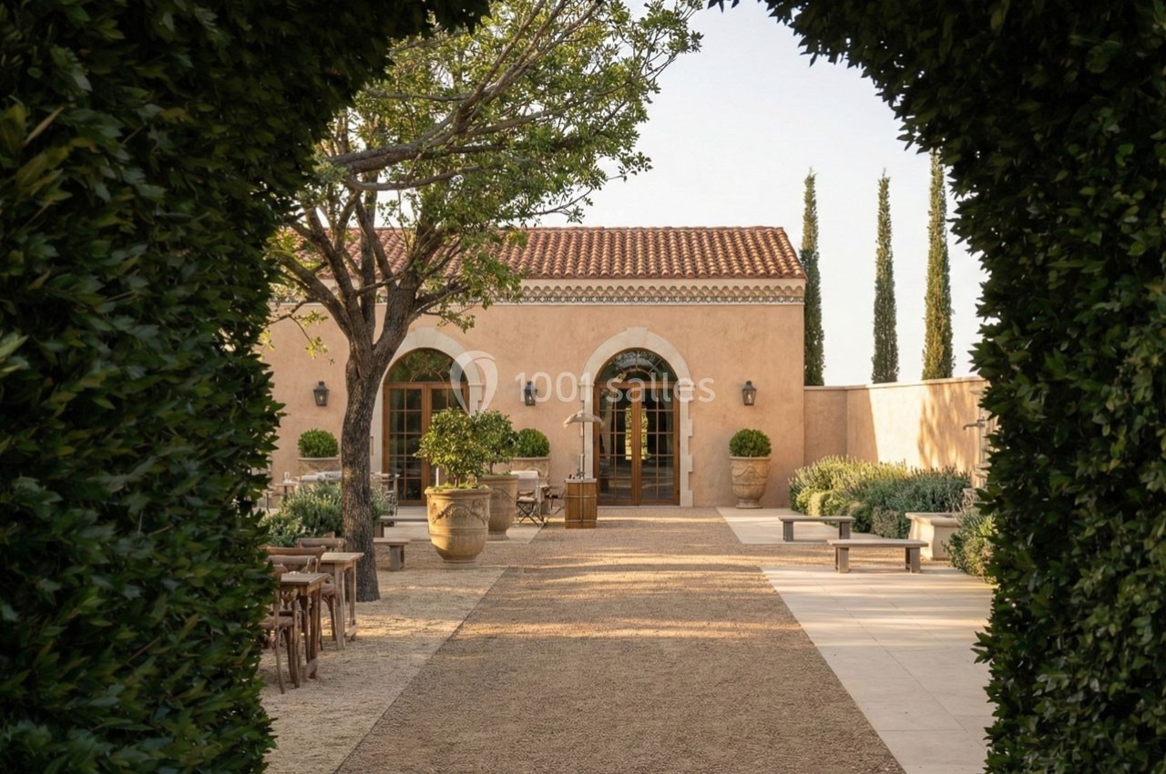 Allée bordée de verdure menant à une bâtisse en pierre avec toit en tuiles, entourée d'arbres et de mobilier extérieur.
