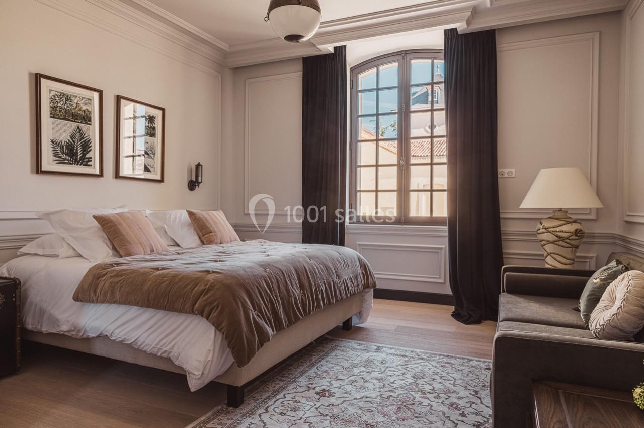 Chambre lumineuse avec un lit double, des coussins beige, un canapé, des rideaux sombres et une grande fenêtre.