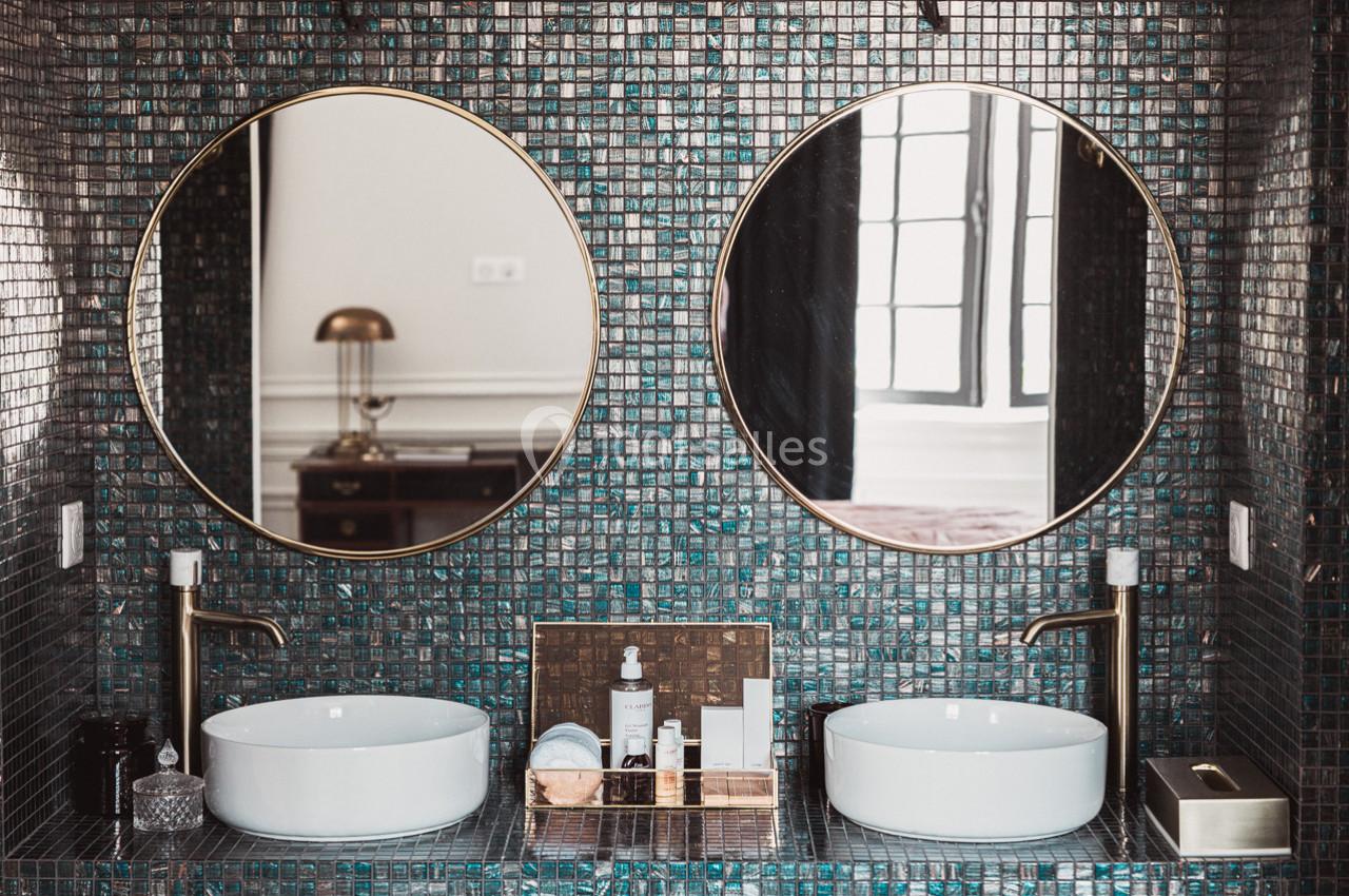 Deux lavabos blancs avec robinets dorés devant un mur en mosaïque sombre, surmontés de miroirs ronds.