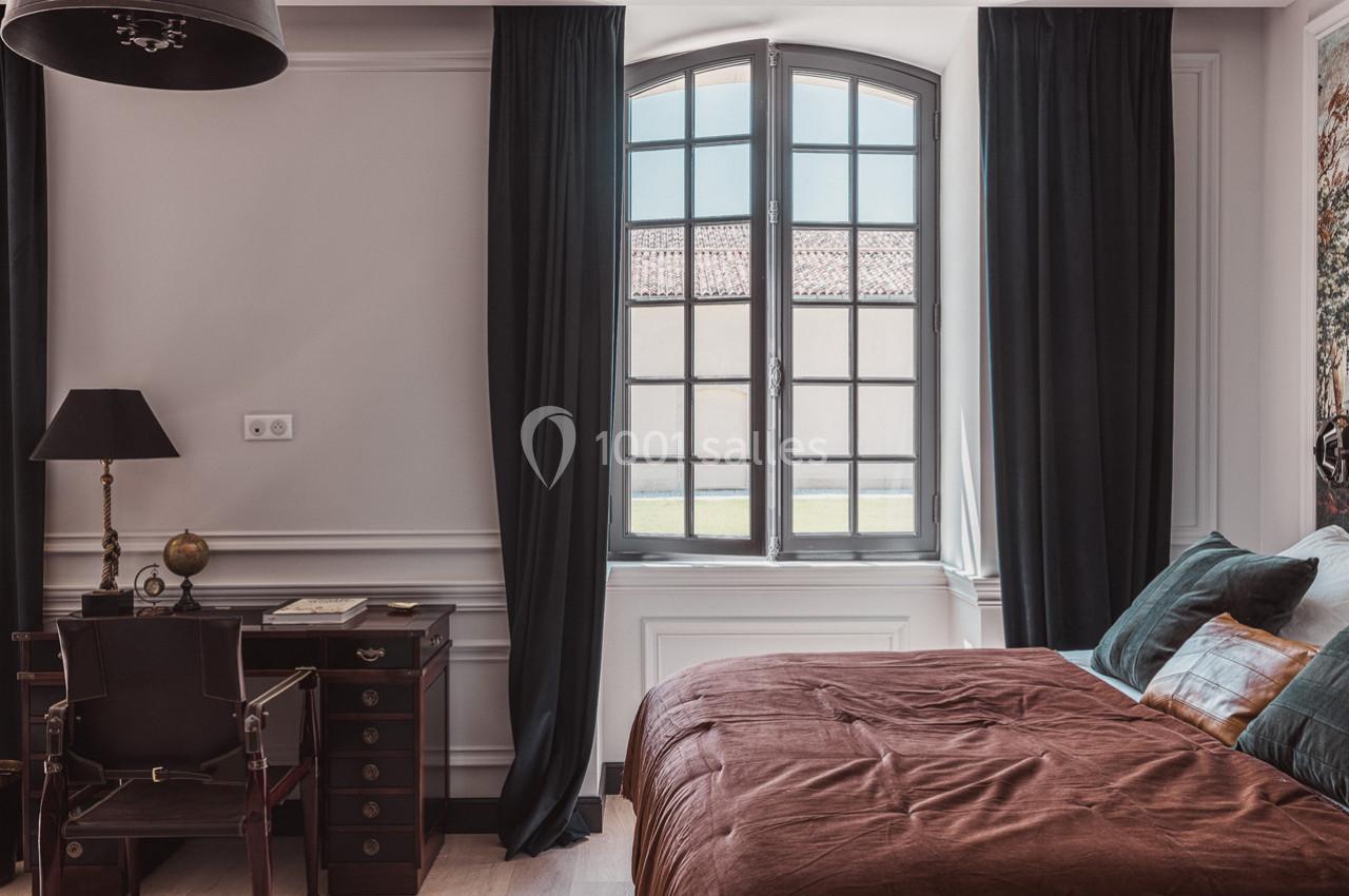 Chambre lumineuse avec un lit, un bureau en bois, une lampe et une grande fenêtre encadrée de rideaux sombres.