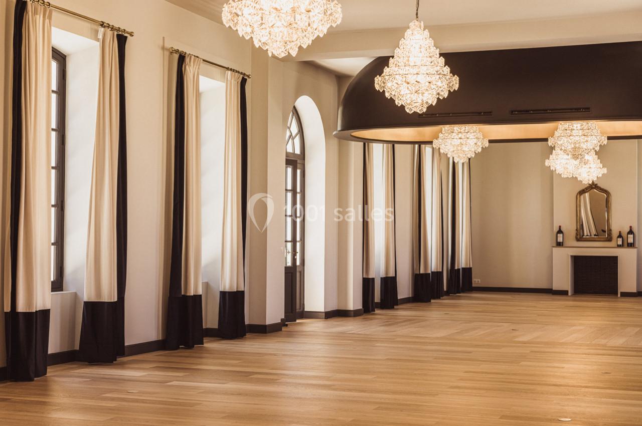 Salle lumineuse avec parquet en bois clair, lustres élégants, grandes fenêtres et rideaux bicolores.