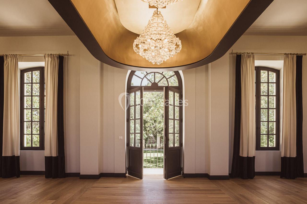 Salle lumineuse avec parquet, lustre central, grandes fenêtres et porte-fenêtre donnant sur un espace extérieur verdoyant.