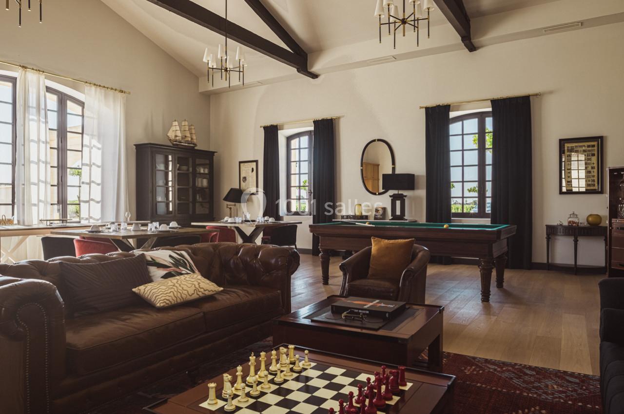 Salle élégante avec canapé en cuir, échiquier, table de billard et grandes fenêtres laissant entrer la lumière naturelle.