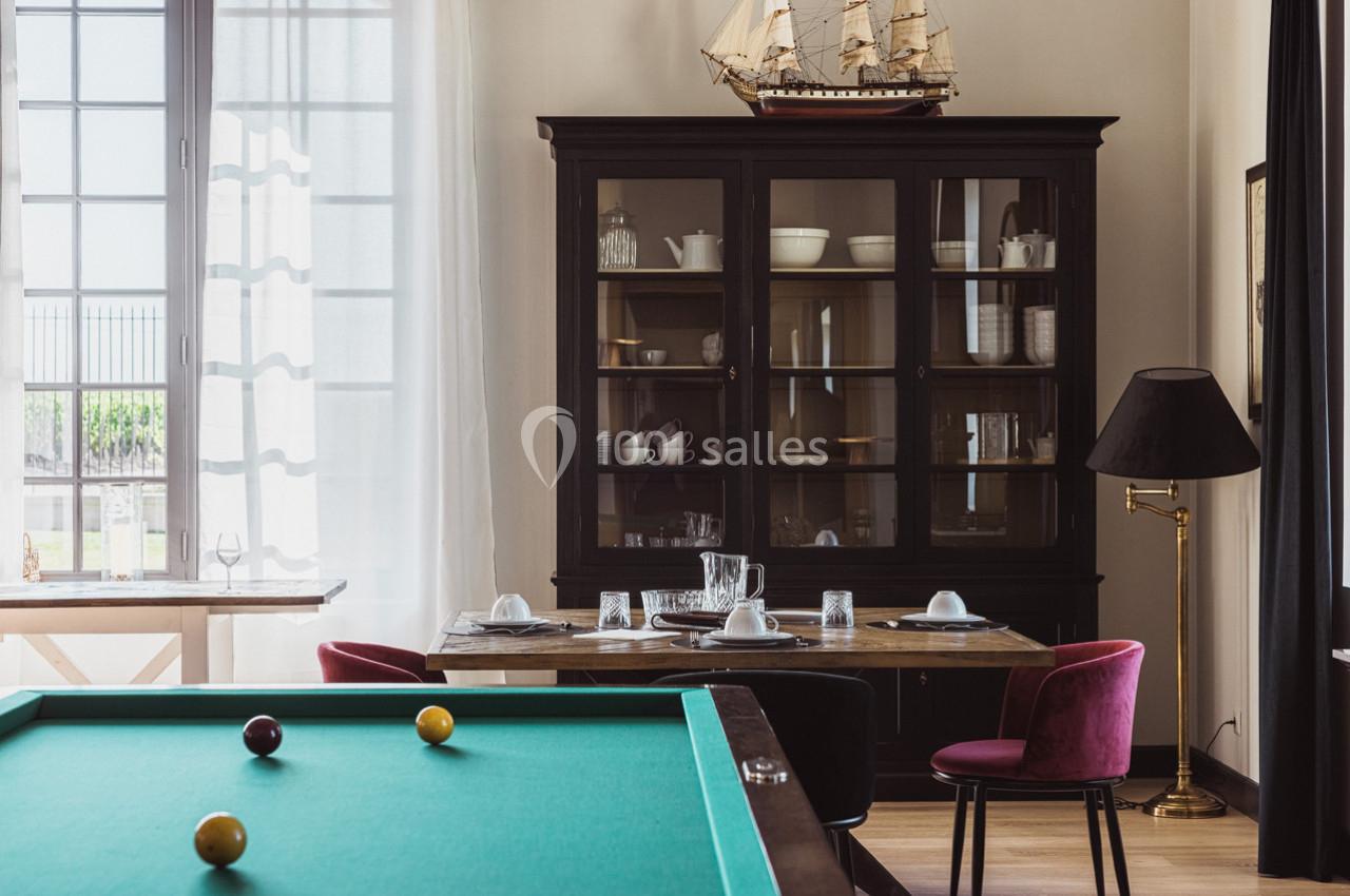 Salle à manger avec table dressée, buffet vitré, lampe sur pied et table de billard au premier plan.