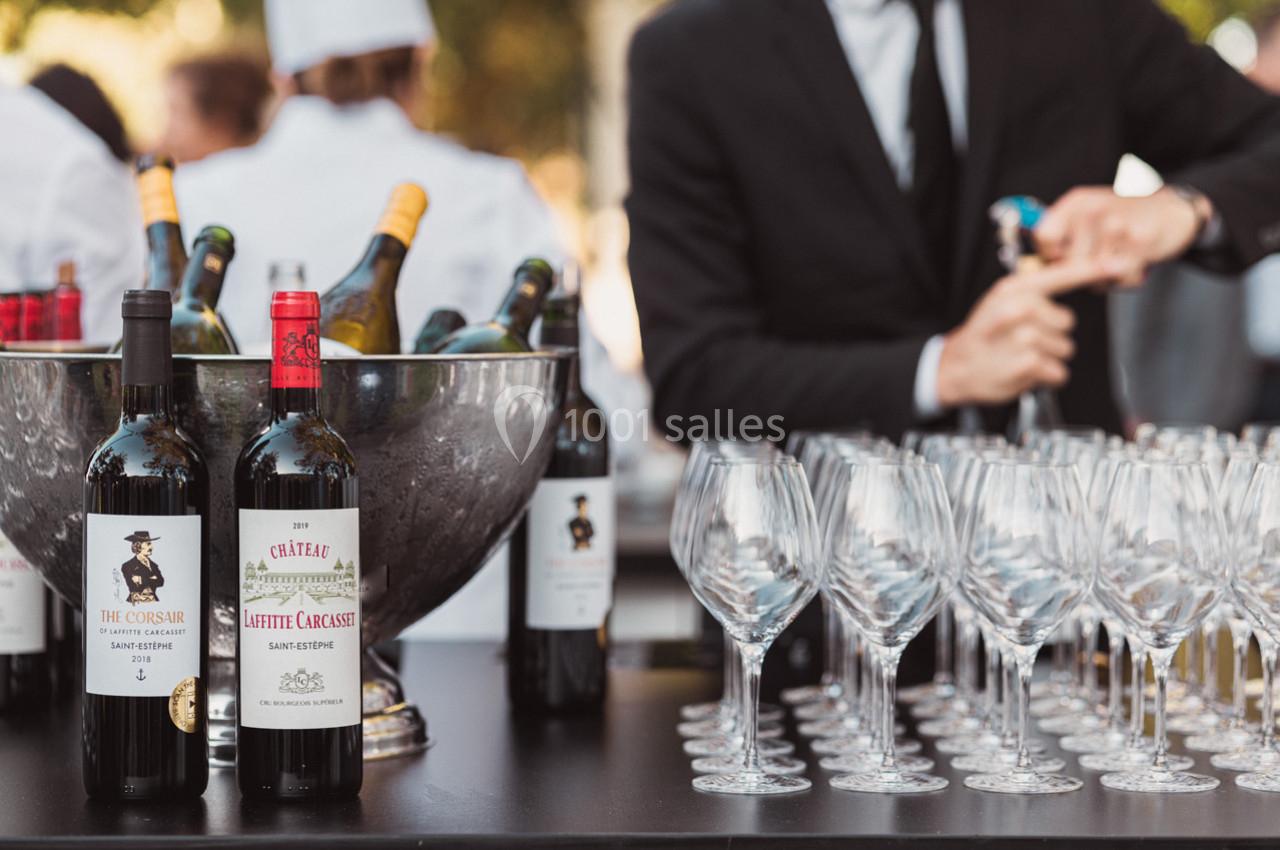 Bouteilles de vin rouge sur une table avec un seau à glace et des verres alignés, un serveur en arrière-plan.